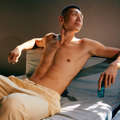 Man sitting on a bed with a bottle of sunscreen in his hand, wearing beige shorts.